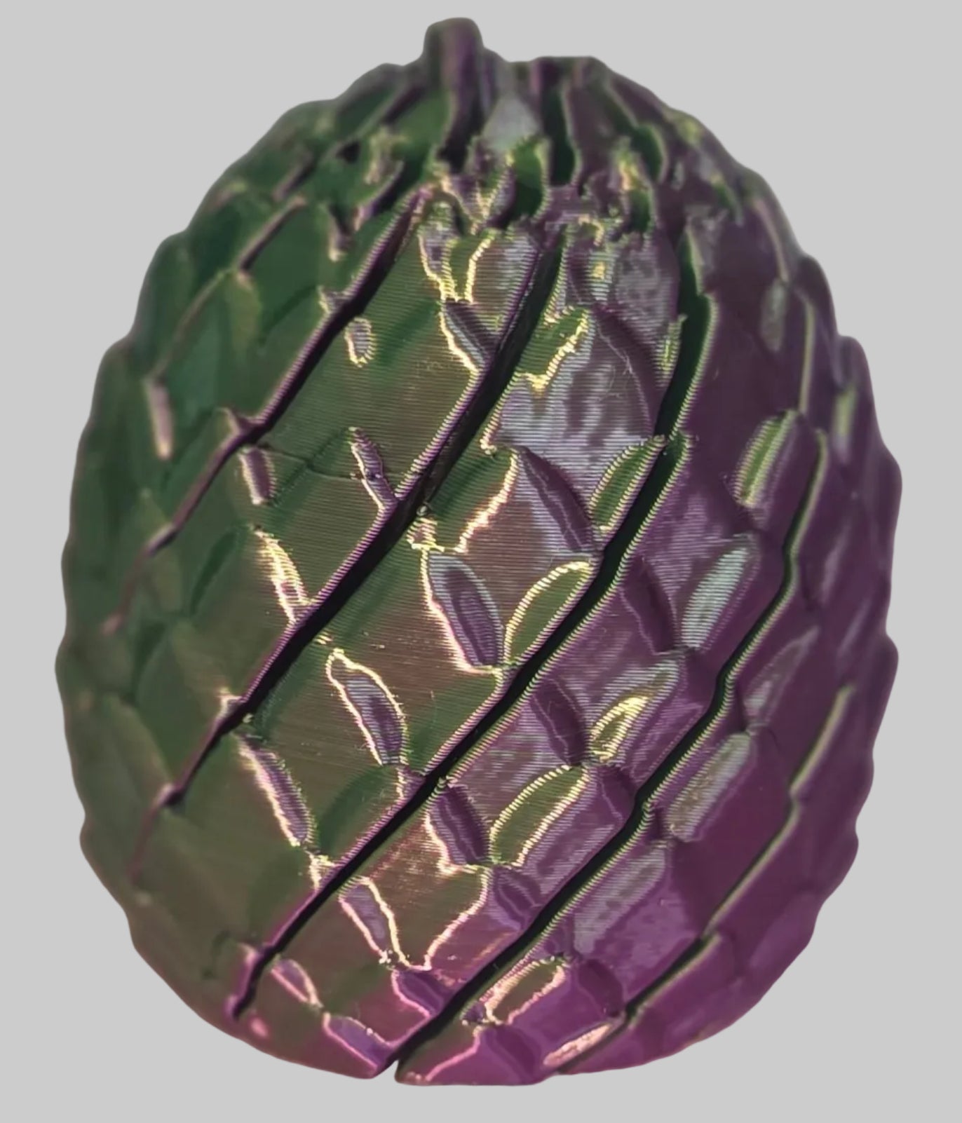 A 3D-printed mechanical egg toy with a textured, segmented surface, shown in a dark green and purple color scheme on a pink surface.
