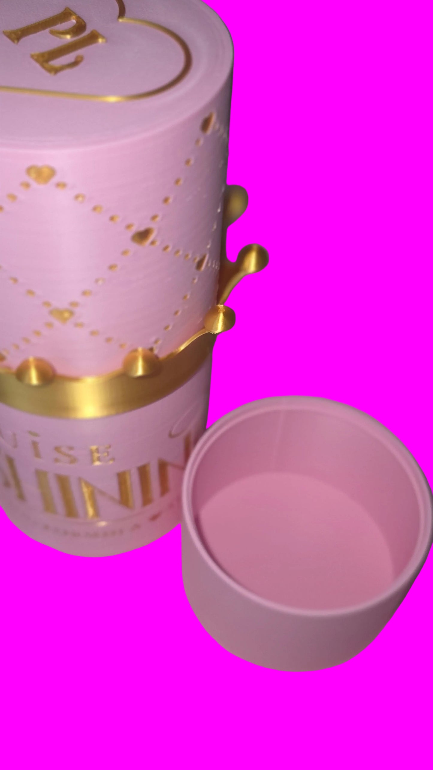 A pink cylindrical cosmetic container with gold decorative lettering and a gold crown motif, shown next to its matching pink lid on a soft pink surface.