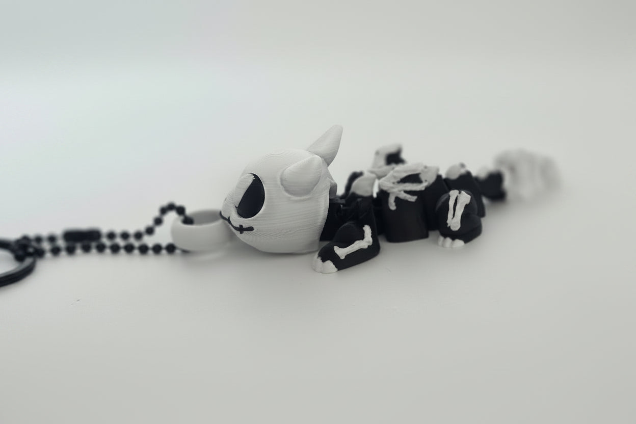 A small plush keychain or pendant shaped like a stylized white creature with black eye details, wearing a black and white skeleton outfit, attached to a black beaded chain on a plain light background.