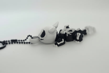 A small plush keychain or pendant shaped like a stylized white creature with black eye details, wearing a black and white skeleton outfit, attached to a black beaded chain on a plain light background.